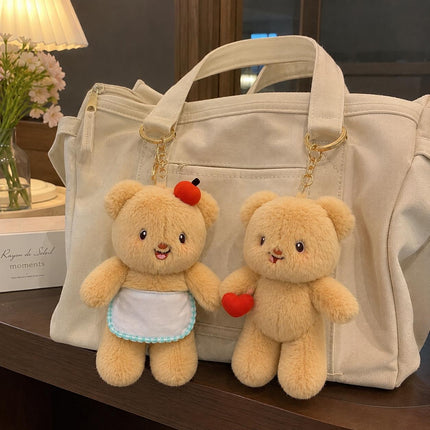 Two light tan teddy bear plush bag charms, one with apple and apron and one holding a red heart, on a handbag